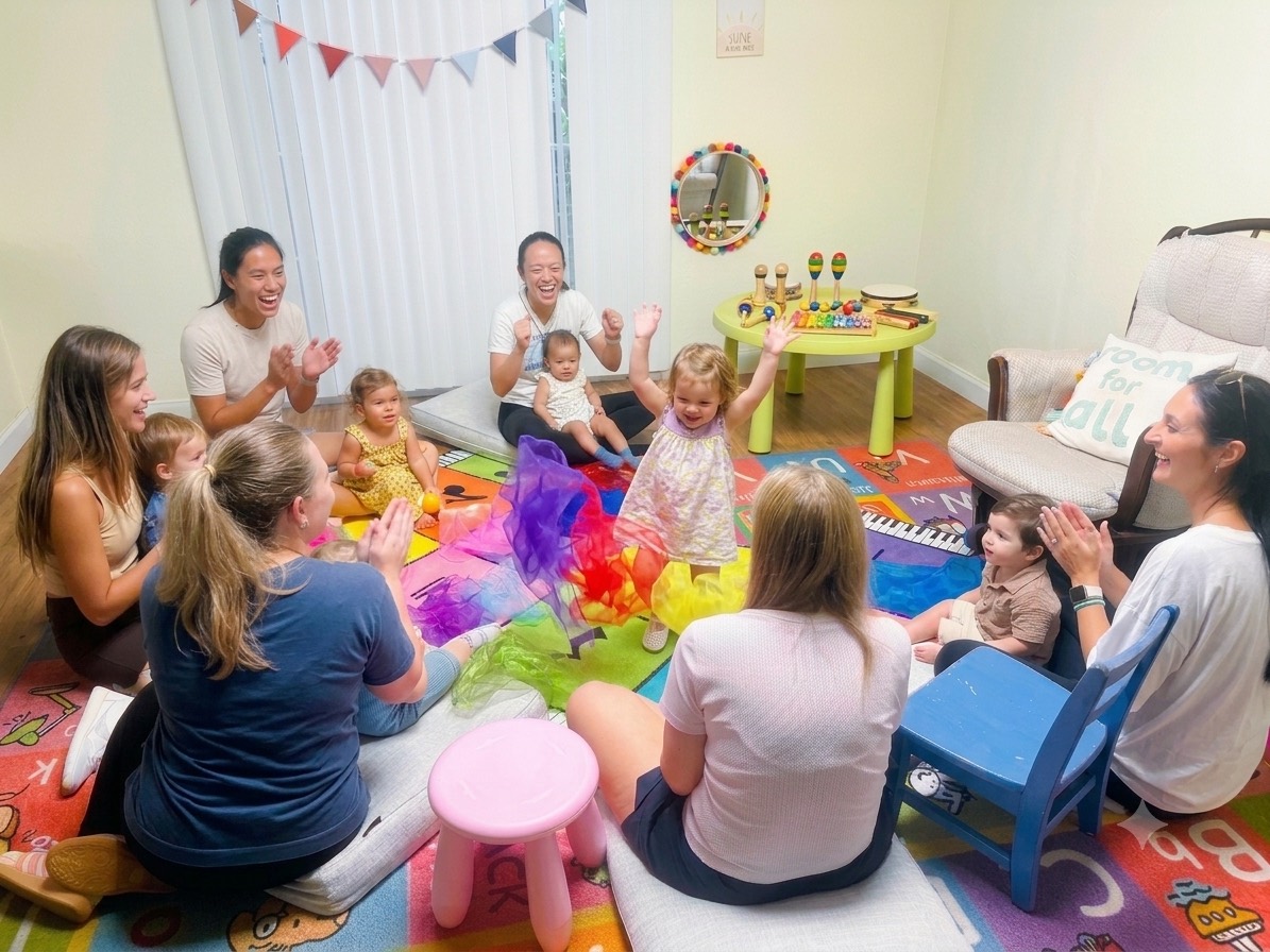 Caregivers clapping as a toddler tosses a scarf overhead during a Mommy & Me music class